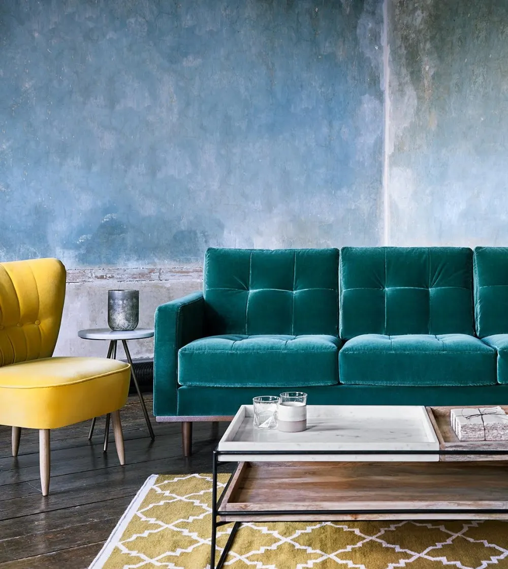 A low rise tufted sofa in teal velvet sits in a next to a mustard occasional chair